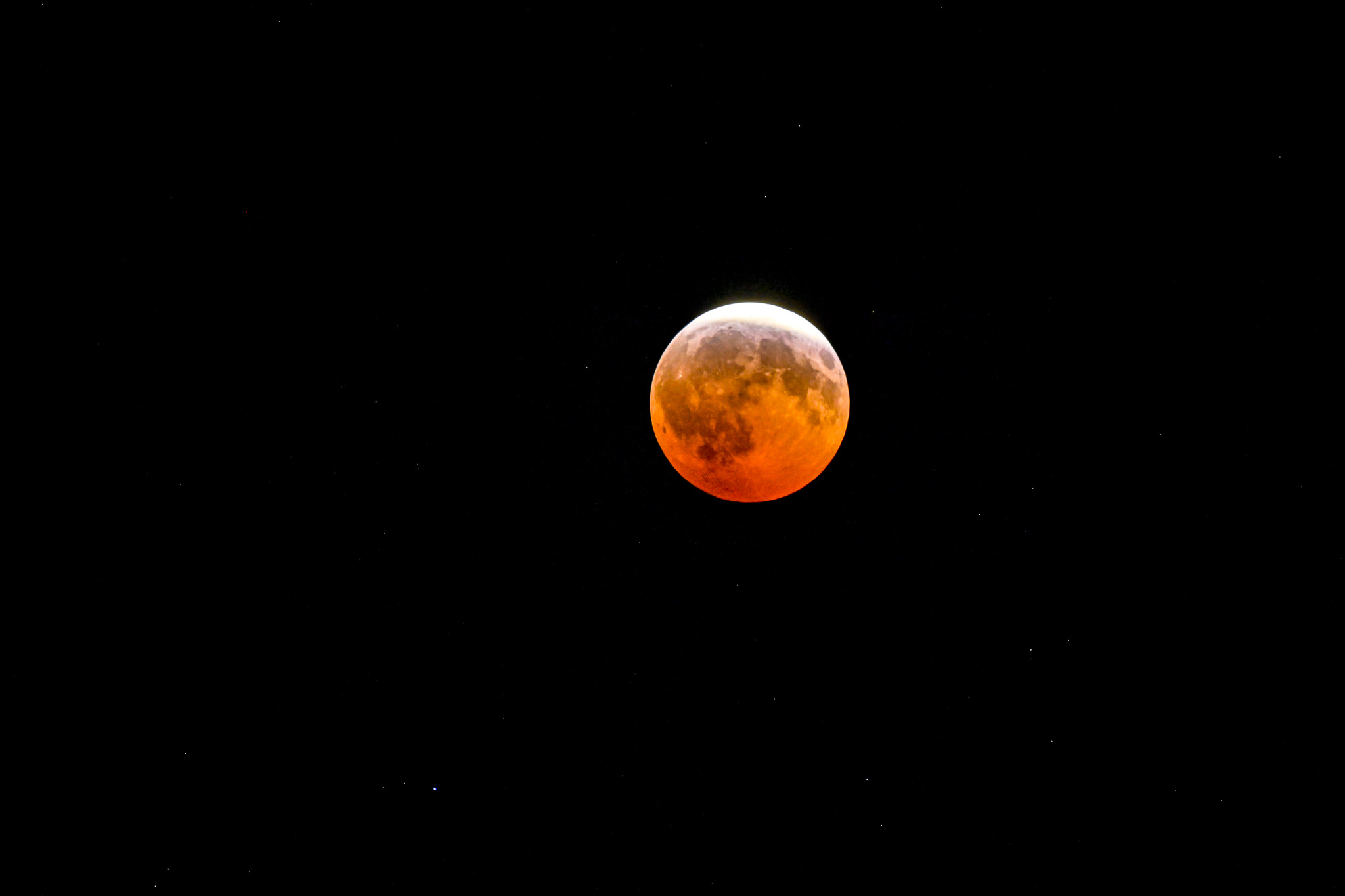 An orange-red eclipsed Moon with a bright upper edge against a starry black sky